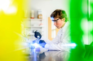 Pedro Graziosi, a Northeastern student in a white lab coat and safety goggles, uses a pipette while working at a lab bench, framed by blurred yellow and green equipment in the foreground.