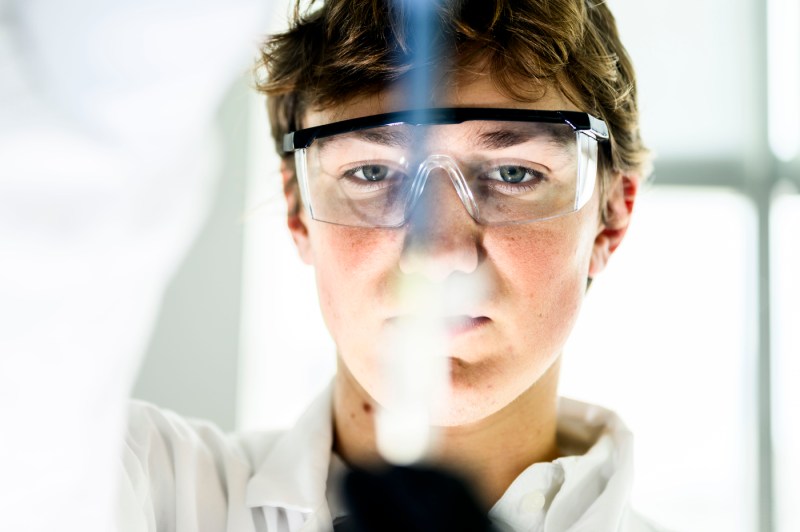 Pedro Graziosi, a biology student wearing safety goggles, looks closely at a test tube in a lab, with the glass creating a blurred vertical line down the center of his face.
