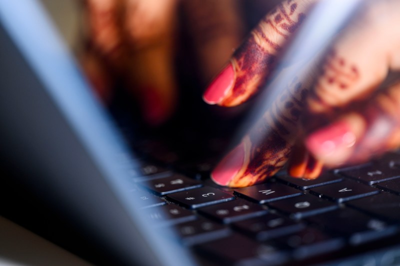 A close up view of Sohni Rais' hands typing on a laptop keyboard. Her fingers are tattooed with henna and her fingernails are pink. 