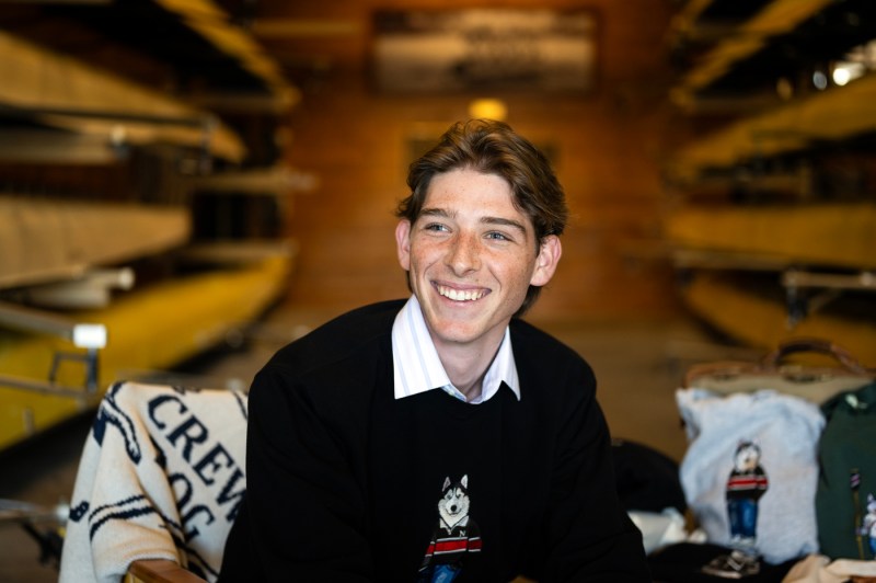 Nicholas Walding smiling and posing for a portrait with custom-embroidered sweaters behind him. 