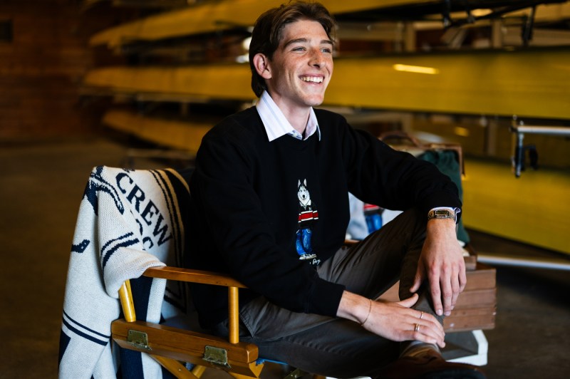 Nicholas Walding sitting in a wooden chair with one foot crossed over one knee. He is smiling and wearing a dark sweater with an embroidered husky on it. 