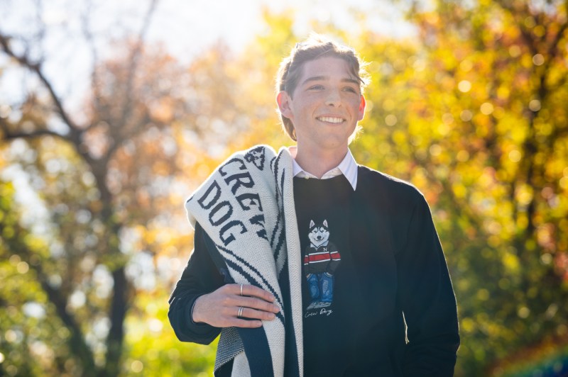 Nicholas Walding standing outside, wearing a sweater with an embroidered husky on it, illuminated by the sunlight. 