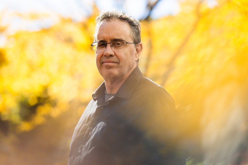 Spyridon Nikas wearing glasses stands outdoors with blurred yellow foliage in the background. 