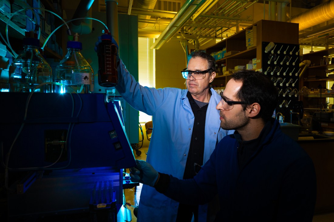 A man in a blue labcoat and goggles stands over the shoulder of another scientist, watching him work. They're next to a table holding flasks and instrumentation.