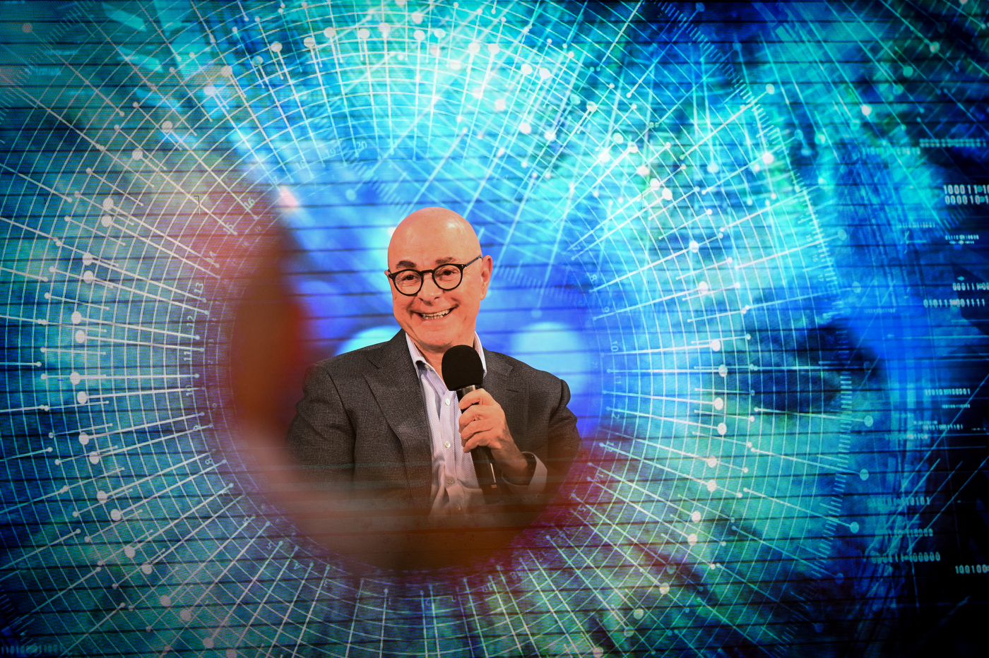 President Aoun wearing round glasses and a gray suit jacket over a light-colored shirt holding a microphone and smiling. A blue and turquoise grid-like geometric pattern is overlaid on the image around him.