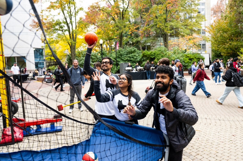 Three students play basketball at a portable hoop during Husky Spirit Day on Snell Quad, surrounded by fall foliage and a crowd of Homecoming Week attendees.