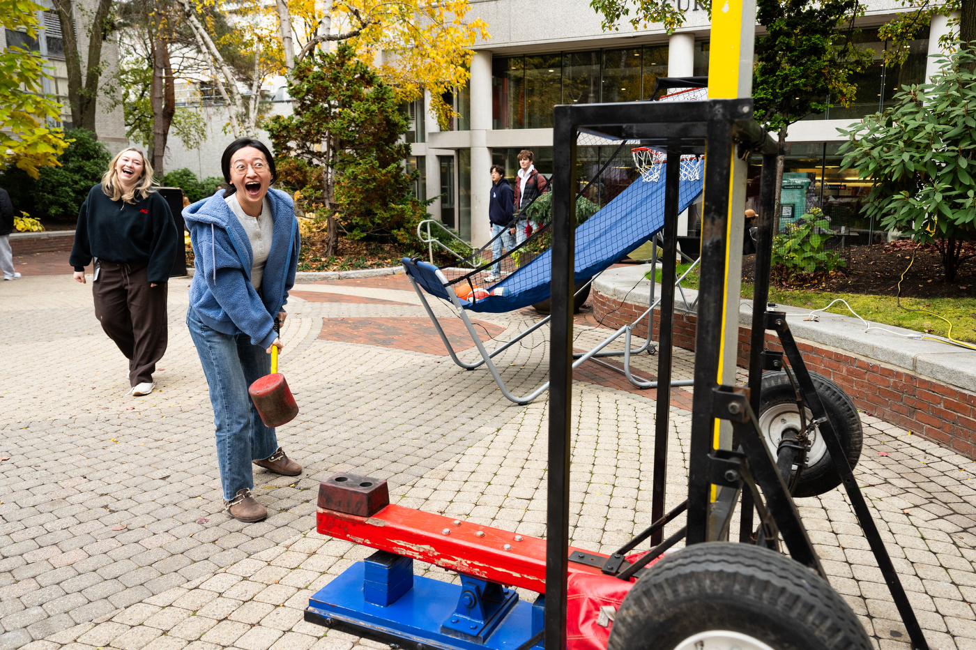 A person wearing a blue hoodie and jeans holding a large hammer in front of a higher strike carnival game that tests the person's strengths. 