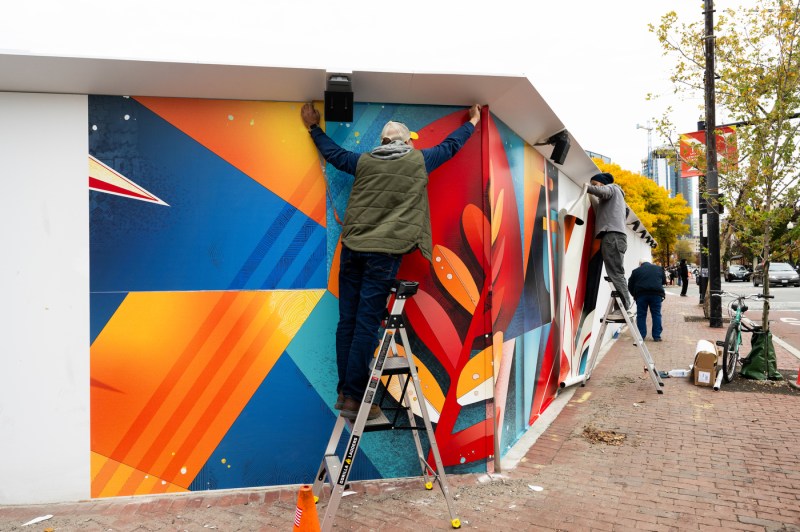 An artist standing on a ladder installs a vinyl art print on a wall on the Boston campus.