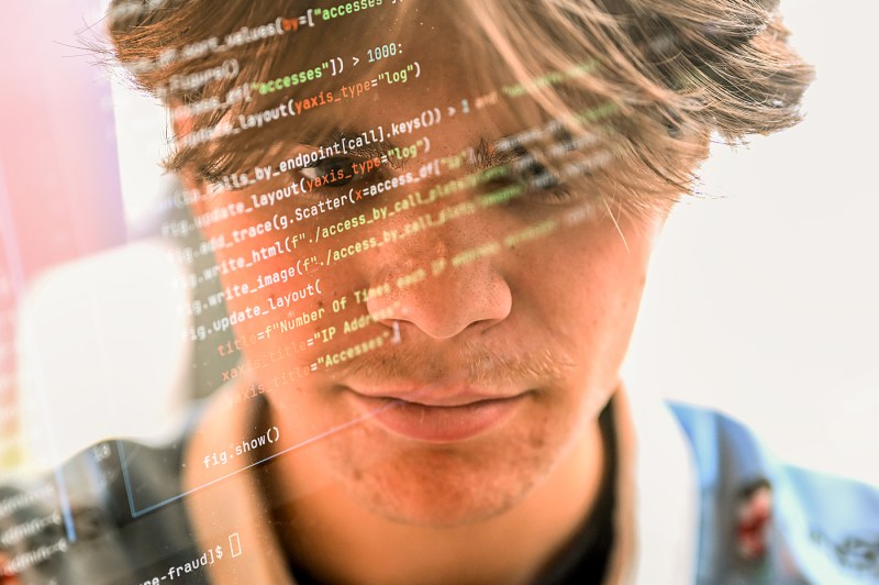 A close-up profile view of Daniel Sheehan, smiling slightly. Lines of programming code are overlaid across the image in orange and green text, creating a technological visual effect.