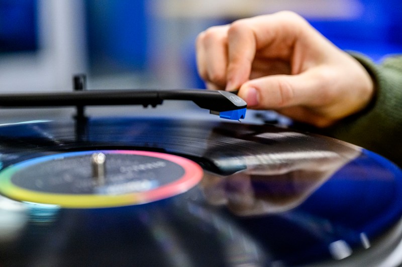 A close-up view of a hand holding a vinyl record over a turntable, with the person's arm extending toward the camera. The background is blurred with blue tones.