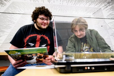 One man places a vinyl record on a turntable while another looks on holding an LP cover