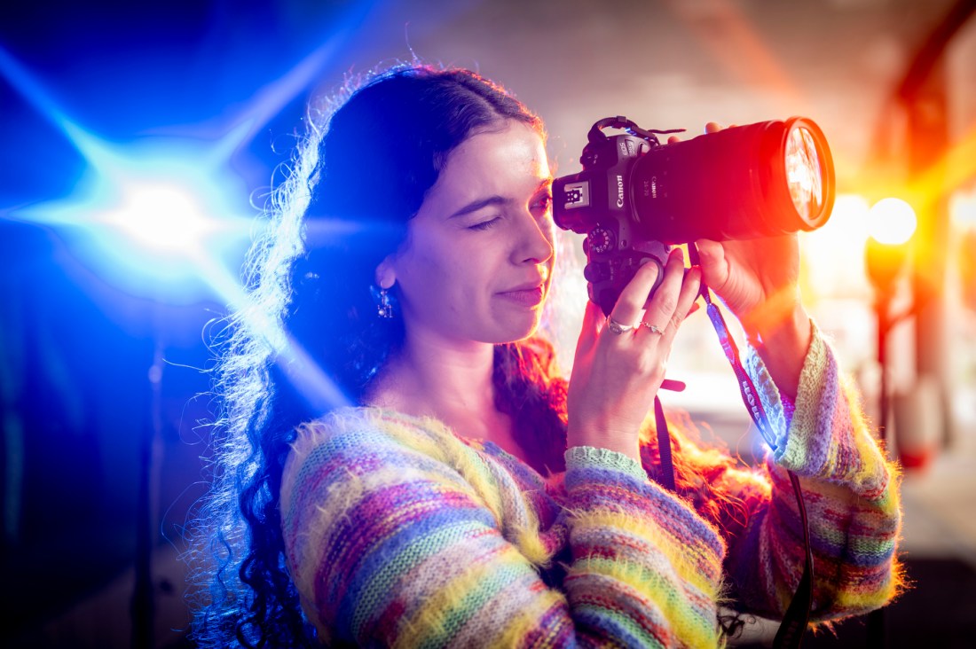 Samantha Davidson closing one eye and looking into the lens of her camera as blue and red lights shine on either side of her.