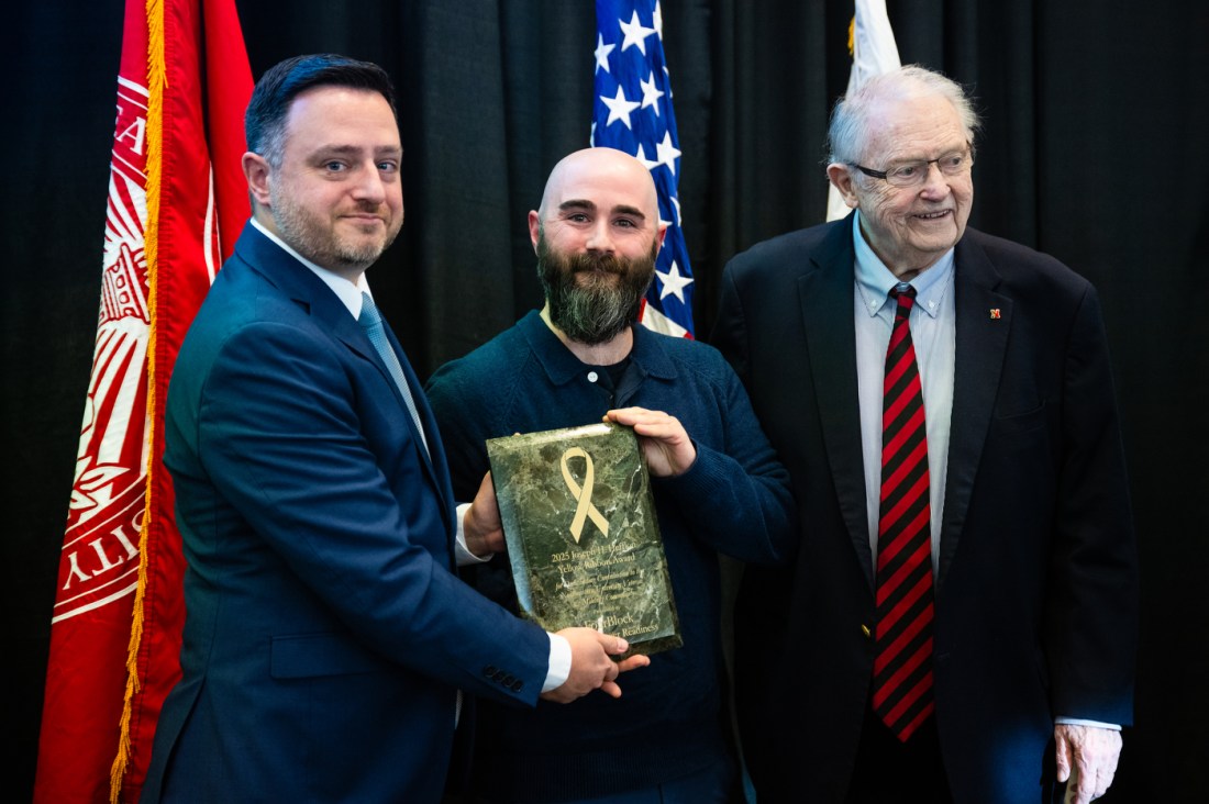 Three people posing together holding a commemorative plaque with a gold ribbon design on it. 