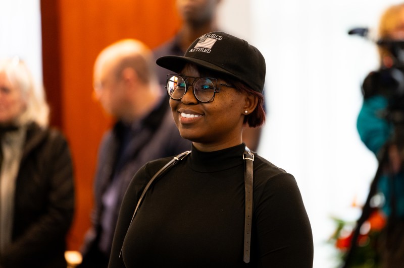 A person wearing a black baseball cap with a US flag embroidered on it smiling and standing at the 2025 Veterans Day Ceremony. 
