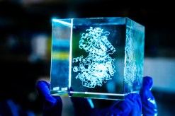 A hand in blue rubber gloves holds a clear plexiglass 3D cubic model of amino acids that look like curly ribbons.