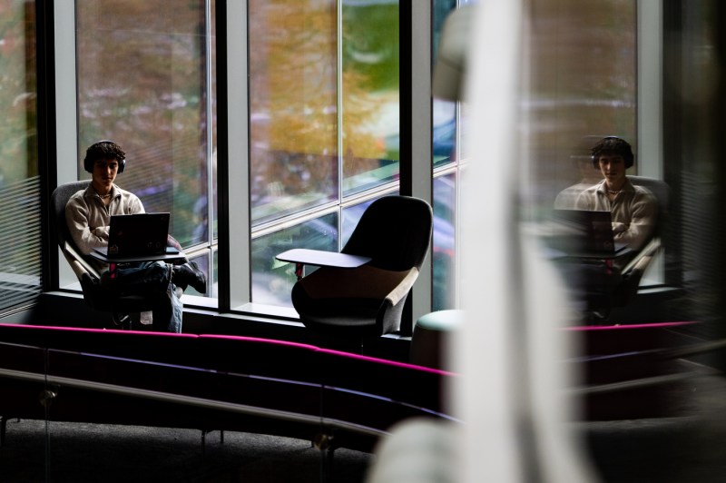 A member of the Northeastern community, who is working on a laptop, is reflected in the glass in ISEC.