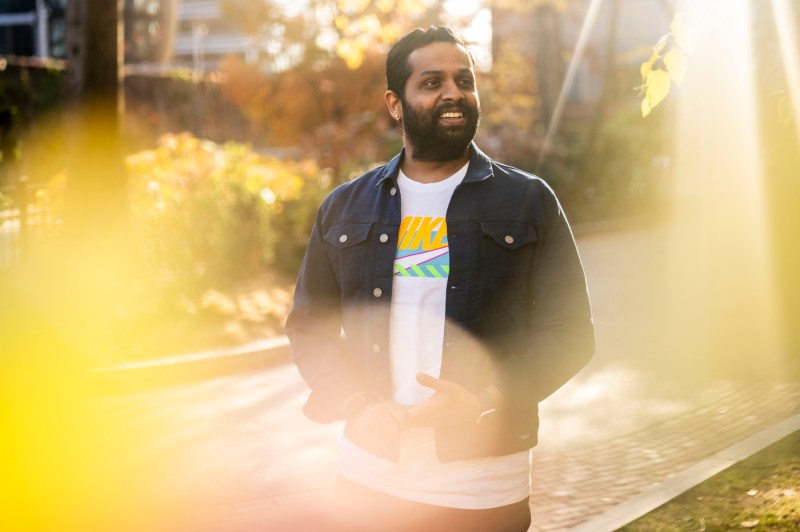 Sundararaman Rengarajan poses for a portrait while standing outside, wearing a white t-shirt with colorful Nike branding on it and a blue jacket over top. 