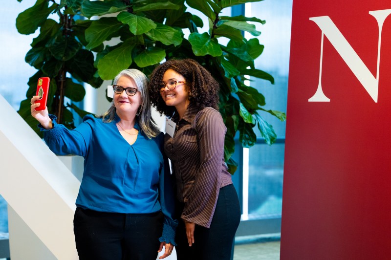 Two women take a selfie in front of a red display with white letter "N" on it.