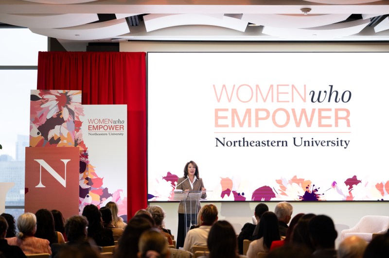 A wide shot of a woman with brown hair in a white-collar shirt and a brown checkered blazer speaking at a clear plastic podium during the event with a white background behind her that says "Women Who Empower Northeastern University."