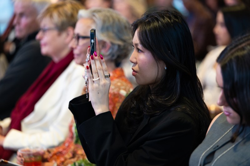 A dark-haired woman in a black blazer holds up her phone to take a photo or video while seated in the audience. She has long red nails and wears several rings.