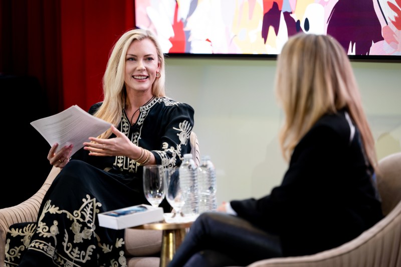 On stage, a blond woman in a long black dress with cream accents sits in a beige swivel armchair as she speaks with another blond woman in a black blazer, who is also seated in a beige swivel armchair.