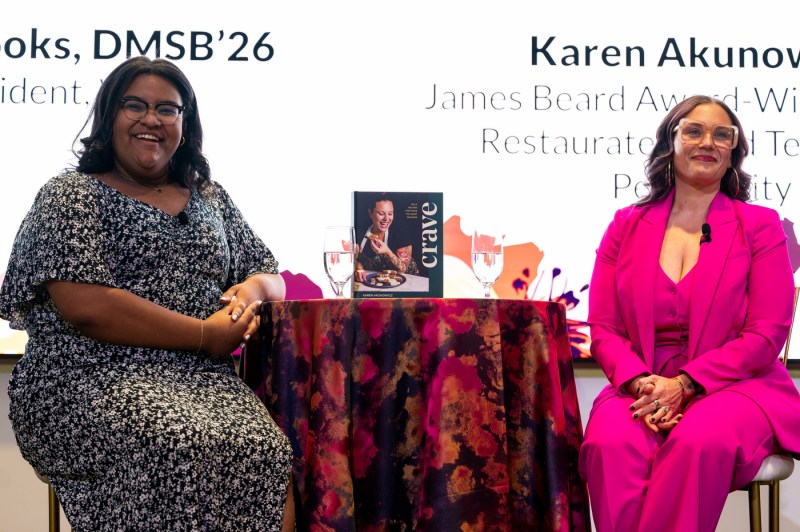 On stage, two women sit on tall chairs with a tall table between them, covered with a brightly patterned floral cloth and two glasses of water on top of it. The woman on the left, wearing a black dress with small white flowers, smiles and leans on the table with one elbow. The woman on the right wears a magenta three-piece pantsuit and light pink glasses.