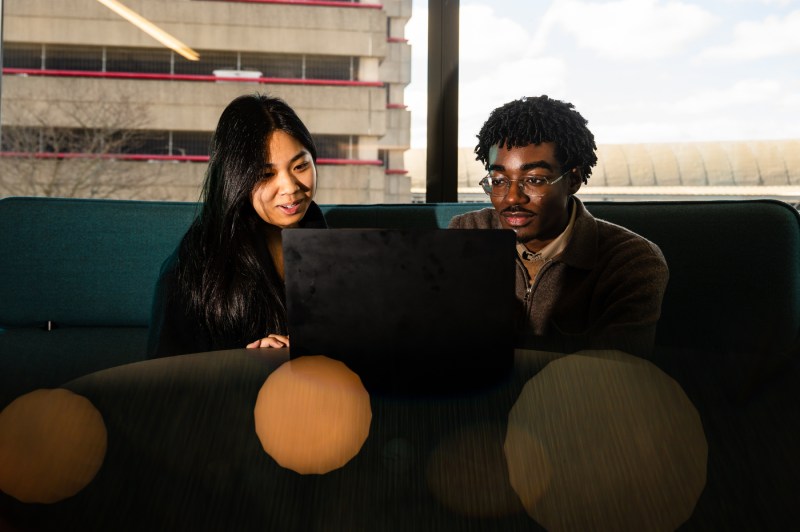 Two students sit on a bench while working on a laptop. There is a window behind them, which overlooks a parking garage.