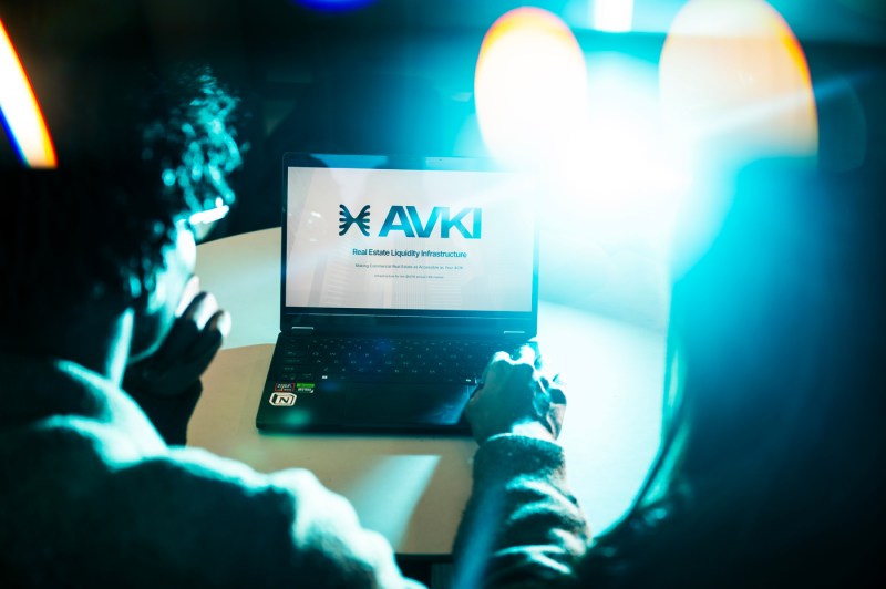 A person, whose back is shown, works on a laptop. The logo for AVKI Real Estate Liquidity Infrastructure is seen on the screen.