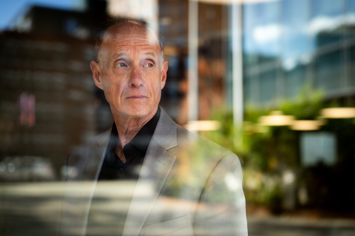 Seen through reflective glass, a man in a grey suit and black shirt looks off camera.