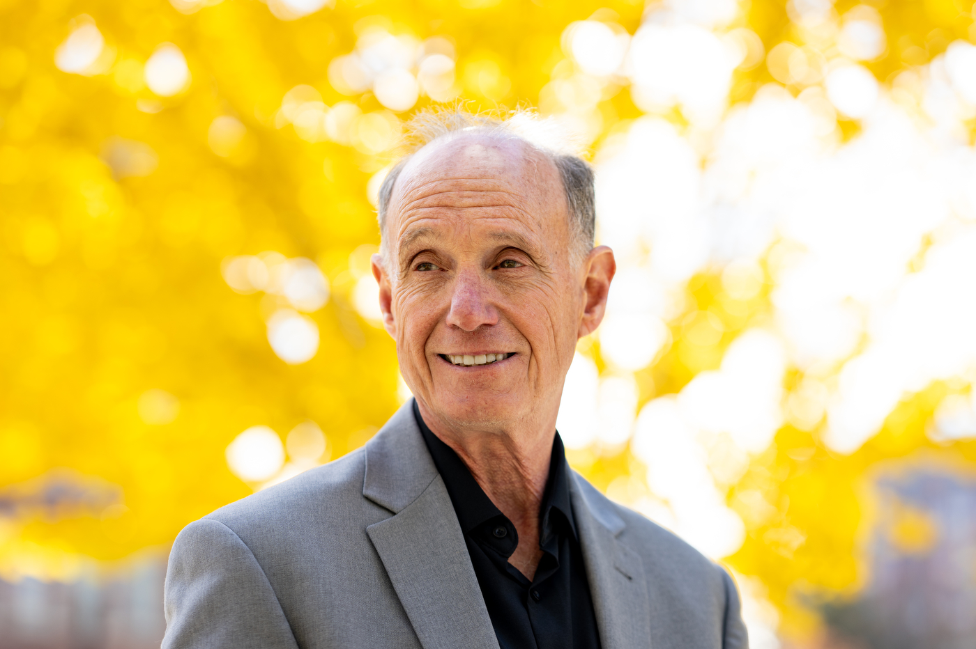A man in a grey suit and black shirt smiles off camera with a bright yellow tree behind him.