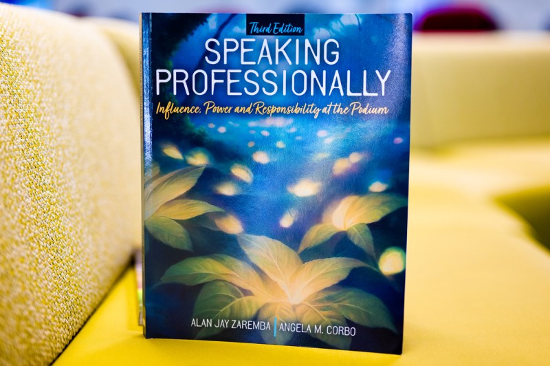 The cover of "Speaking Professionally," predominantly blue with yellow flowers in the foreground. The book rests on a yellow couch.