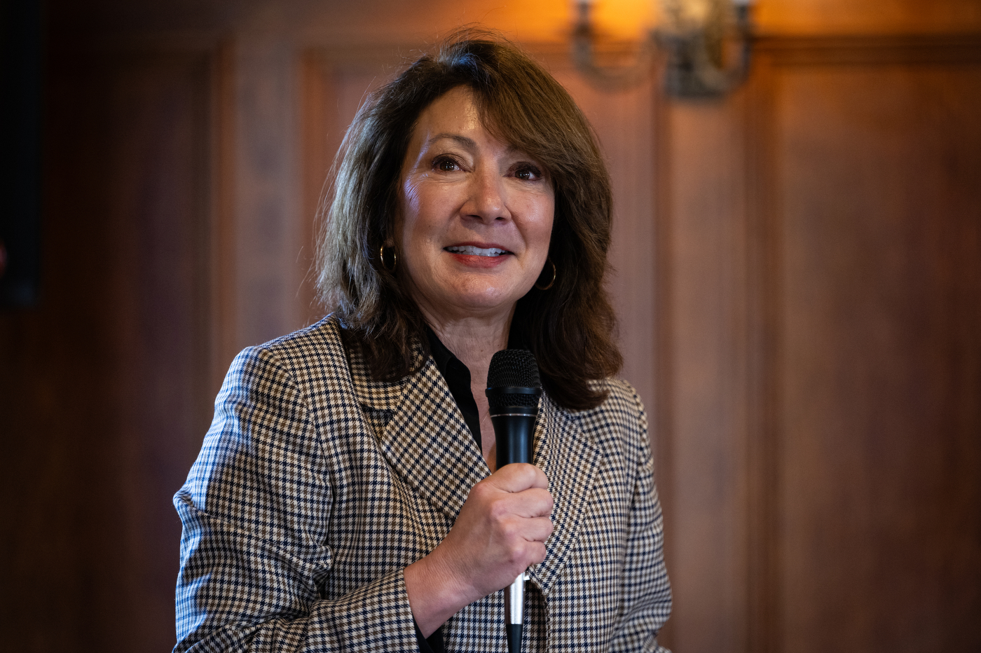 Diane MacGillivray, a woman with shoulder-length brown hair, smiles while seated in a warmly lit wood-paneled room during a formal event.