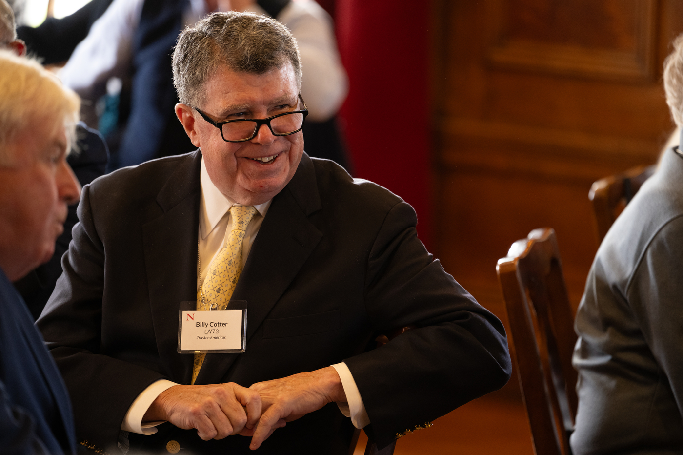 A man with a yellow tie and glasses smiles while listening to a speaker at the luncheon event.