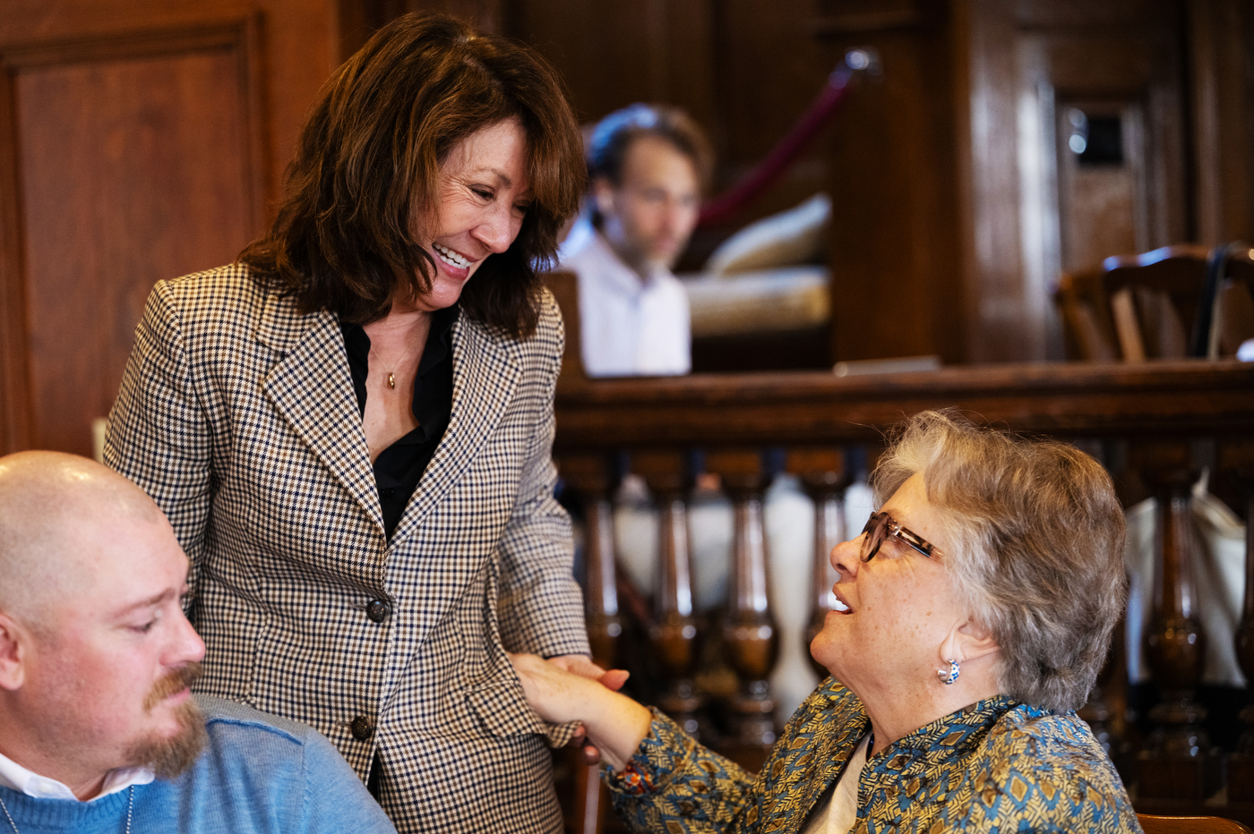 A woman with brown hair smiles and looks down while seated at a table in a warm, wood-paneled room with other attendees in the background.