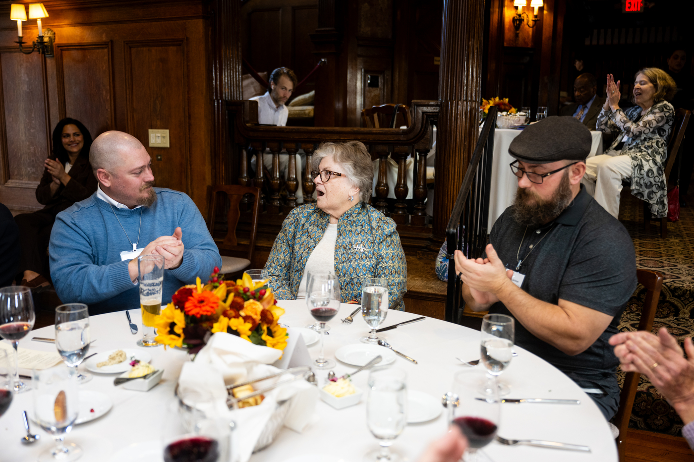 Three attendees sit together at a dining table in a wood-paneled room; one person in a blue sweater gestures while speaking to an older woman, and another person wearing glasses and a cap looks down at their plate.