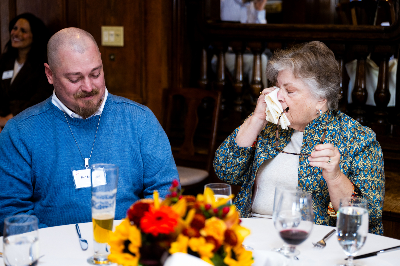A man in a blue sweater smiles while seated next to an older woman who wipes her eyes with a tissue at a dining table, suggesting an emotional moment.
