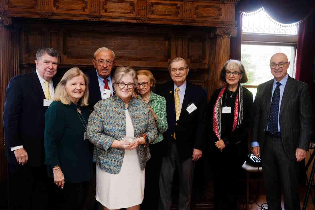 A group of older men and women in suits and blazers gather in front a wooden fireplace.
