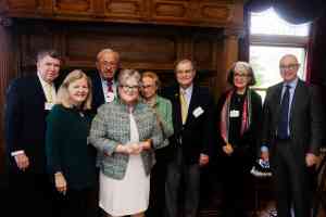 A group of older men and women in suits and blazers gather in front a wooden fireplace.