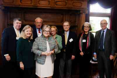 A group of older men and women in suits and blazers gather in front a wooden fireplace.