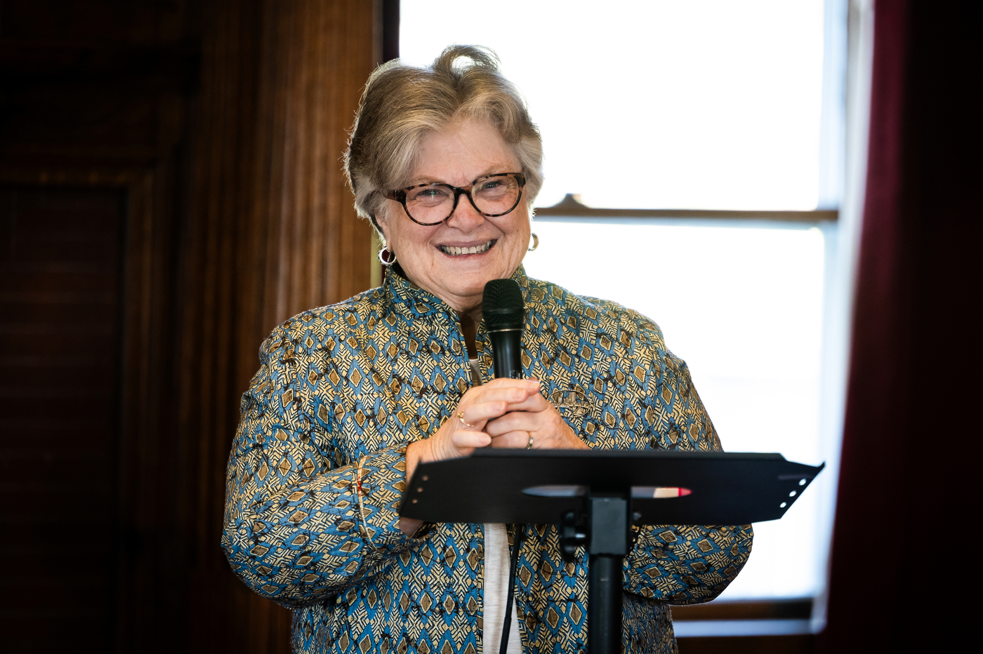 A woman wearing glasses and a patterned jacket smiles while speaking into a microphone at the event.