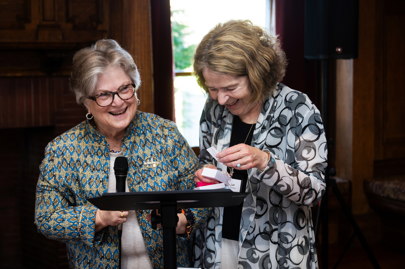 Two women sit side by side at a table, smiling and looking down as one of them adjusts something in her hands.