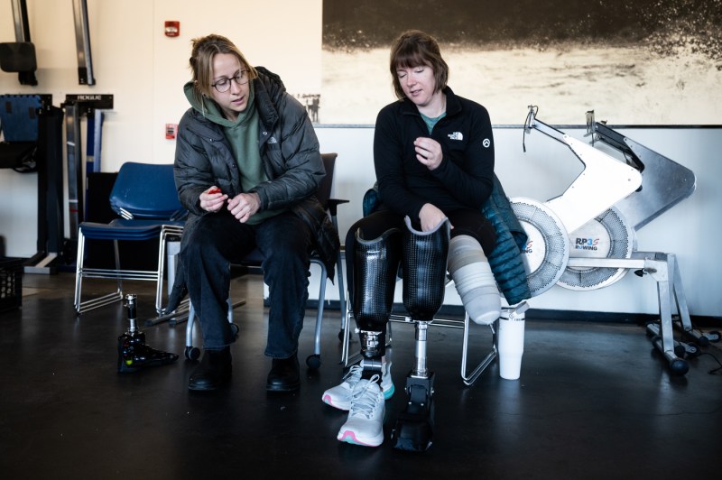 Two people sit together in an indoor rowing facility, one on a chair and one on a rowing machine with a white prosthetic foot and ankle visible, discussing adjustments in a well-lit training space.