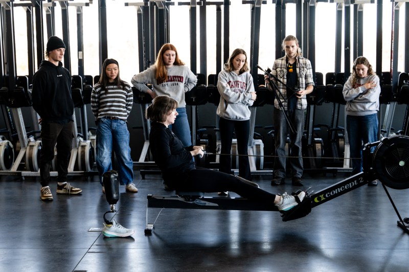 Several students observe as a person tests a prosthetic device while using a rowing ergometer in an indoor training facility.