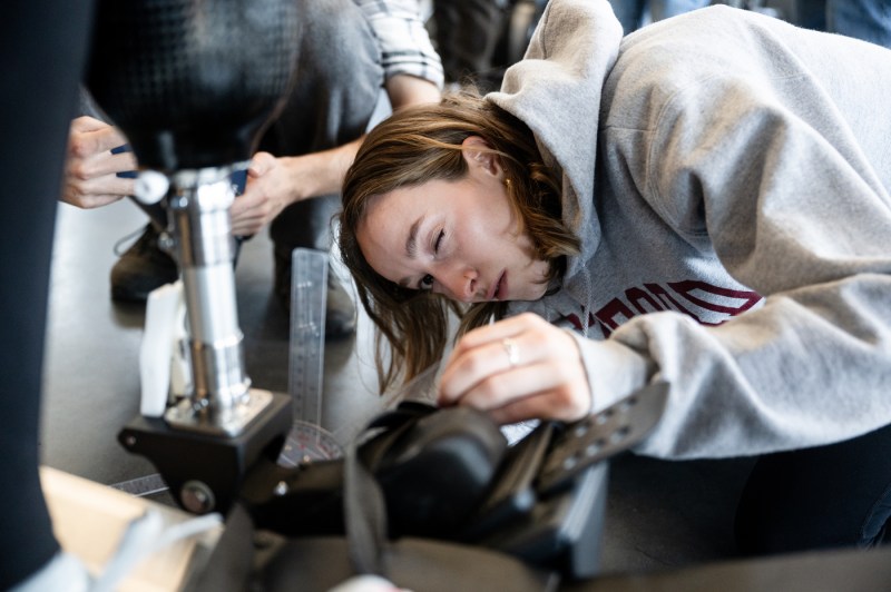 A person in a gray hoodie leans over to examine or work on rowing equipment, with others visible in the background