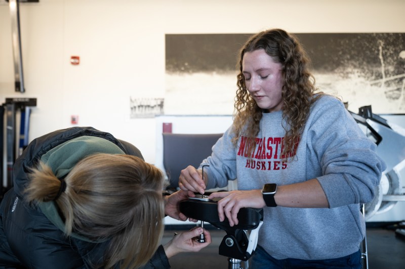 A student in a Northeastern Huskies sweatshirt adjusts rowing equipment while another person with blonde hair looks on in an indoor training facility.