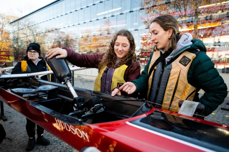 Three people examine adaptive rowing equipment mounted on a red rowing shell, wearing life jackets at an outdoor waterfront location.