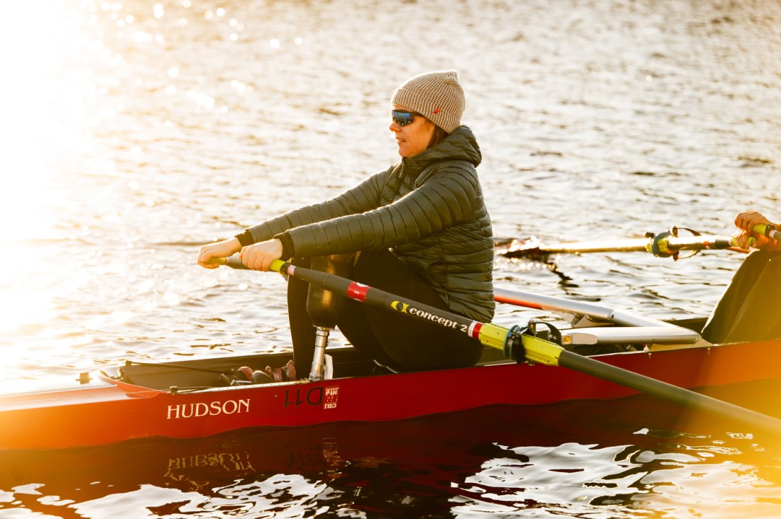 A rower in a tan beanie and sunglasses rows a single scull on calm water, seated in a red Hudson racing shell with Concept2 oars.