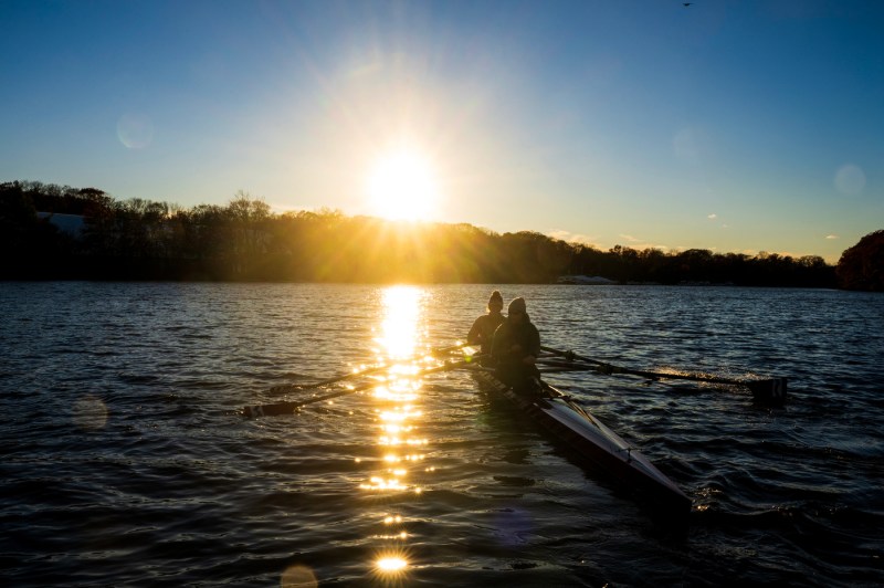 Two people row together in a boat on calm water during sunrise or sunset, with bright sunlight creating a glowing backdrop on the horizon.