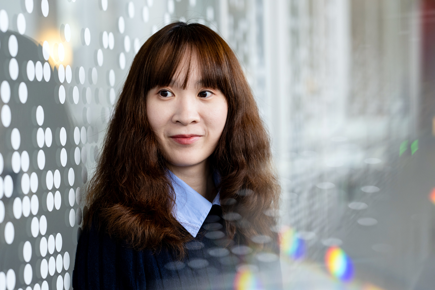 A woman with shoulder-length, dark hair stands in front of a gray background with white dots. Her shirt has a blue collar and she looks off to the left.