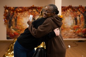 Two women embrace warmly while one holds papers, standing in front of an autumn-themed painted backdrop featuring orange and gold fall trees and pumpkins.
